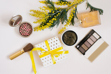 A workspace with yellow mimosa flowers, decorative cosmetics, a gift box with a bow on a white background. Flat lay. Top view female blogger background