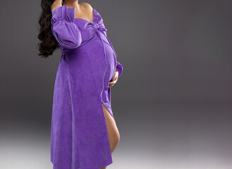 A pregnant woman with long dark hair in a purple dress holds her belly. Pregnancy. Motherhood. A pregnant woman on a gray background with a place for text.