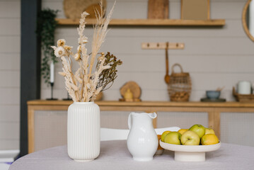 Autumn decor of the dining table. Dried flowers in a white ceramic vase, a jug of milk and apples on a tray or plate in Scandinavian cuisine