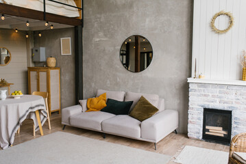 Gray sofa with pillows, a round mirror hangs on the wall in a modern Scandinavian-style living room near the fireplace and dining table