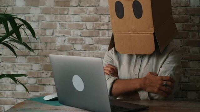 One person with cardboard box carton ho his head without expression working on laptop at the desk in home office. Concept of identity and online internet security business. Catfish cybercrime web
