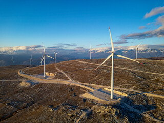 Windmills on the hills during sunset. Renewable energy, green energy. Mountains in the background with snow. Wind power and environmentally friendly. Sustainable future. End fossil fuels.