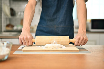 Young man in apron kneading dough with a rolling pin on kitchen table, preparing homemade pastry in kitchen