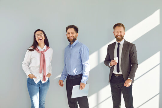 Happy positive cheerful business team having fun during a break at work. Group of three people having a coffee break, standing by a grey office wall, looking at the camera and smiling