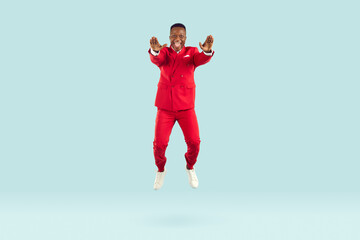 Fototapeta na wymiar Funny stylish african american man having fun bouncing on pastel light blue background. Millennial man in red suit with cheerful crazy expression jumps with his arms straight. Full length. Banner.