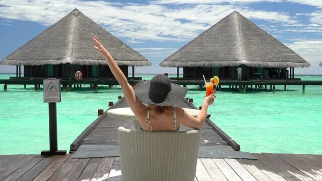 A young happy girl in a big black beach hat sits at a table and holds a bright yellow ceta cocktail with a pineapple heart in her hands against the backdrop of water villas in the Maldives. The girl t