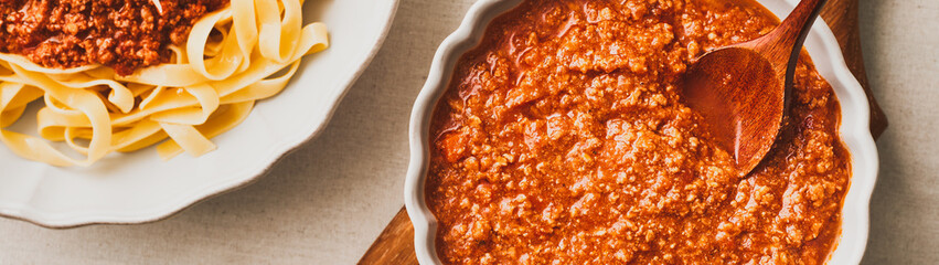 A white bowl of fresh bolognese sauce and a plate of fresh tagliatelle pasta with bolognese sauce. Web banner with copy space. Top view.