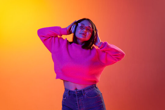 Relaxation. Young girl in cozy sweater listening to music in headphones isolated over gradient orange background in neon light. Concept of emotions, facial expression, youth, inspiration, sales, ad