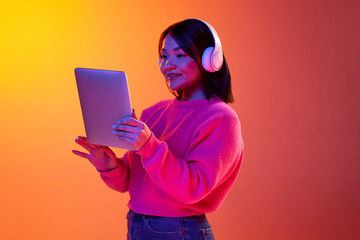 Online lessons. Young girl, student in headphones looking on tablet over gradient orange background...