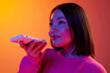 Young girl in pink sweater recording voice message with phone over gradient orange background in...