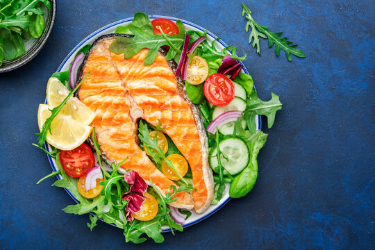 Grilled Salmon Steak With Vegetables, Tomatoes, Arugula And Lettuce Side Dish Salad On Plate, Blue Table Background, Top View