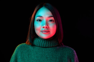 Looking at camera. Young beautiful girl with short brunette hair in cozy sweater over dark background in neon light. Concept of emotions, facial expression, youth, inspiration, sales, ad