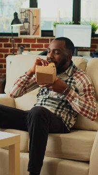Vertical video: Modern man eating hamburger with fries on couch, having dinner with fast food and beer bottle at home. Young guy feeling relaxed watching comedy or action movie, having fun with tv.