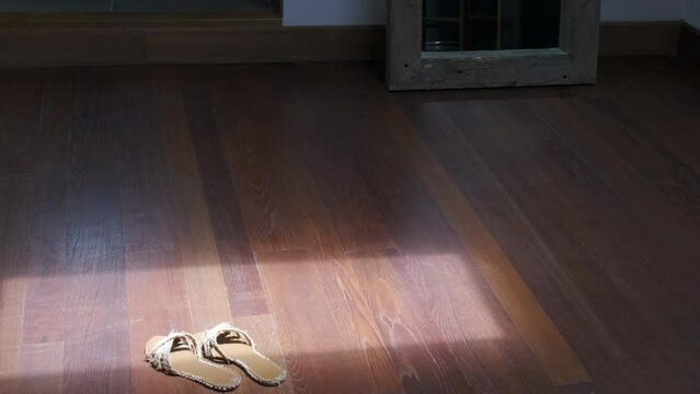 Mujer vestida de blanco se descalza y deja sus sandalias en el haz de luz de una ventana sobre suelo de madera