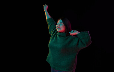 Excitement. Cheerful mood. Young emotional brunette girl in cozy sweater posing over dark background in neon light. Concept of emotions, facial expression, youth, inspiration, sales, ad