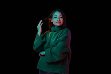 Coziness, comfort. Young beautiful brunette girl in cozy sweater posing over dark background in neon light. Concept of emotions, facial expression, youth, inspiration, sales, lifestyle, ad