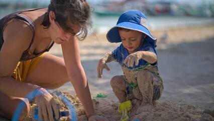 Cute little boy covered in sand playing with his auntie on the beach with toys on a sunny afternoon