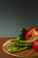 Vegetables lie on a wooden board: tomatoes, asparagus, cucumbers, red bell peppers. brown, dark gray background. place for text.