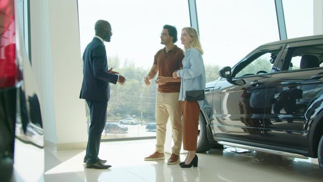 Long shot of African American man working in dealership center giving car key to customers