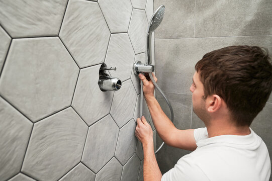 Male Worker Fixing Metal Bath Showerhead And Doing Plumbing Works While Working On Home Renovation. Man Standing By The Wall With Ceramic Tile And Installing Handheld Showerhead In Bathroom.