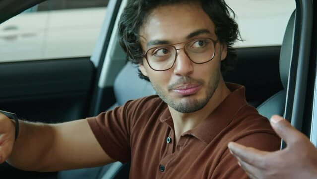 Joyful young Middle Eastern man sitting in car in dealership showroom discussing technical specifications of crossover model with salesperson