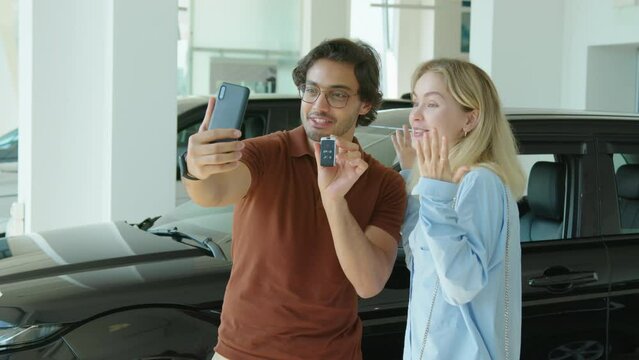 Modern young couple standing in dealership showroom holding smartphone recording video about buying new car for blog