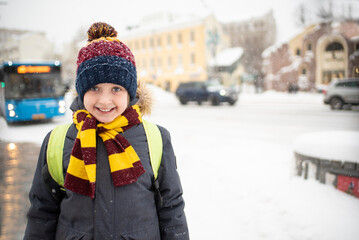 A smiling boy, a schoolboy, a child in a hat and jacket stands at a bus stop, waiting for a public...