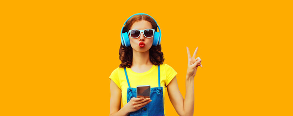 Portrait of stylish young woman in headphones listening to music with smartphone on yellow...