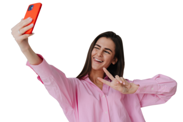 Excited brunette girl in pink shirt winks, makes selfie at transparent background. Satisfied blogger during travel tour. Cheerful student holds phone making video call shows peace gesture, tongue out.