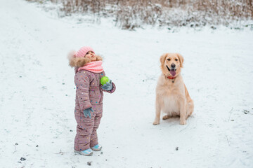 Young girl portrait in pink hat playing active game with her dog golden retriever in winter season