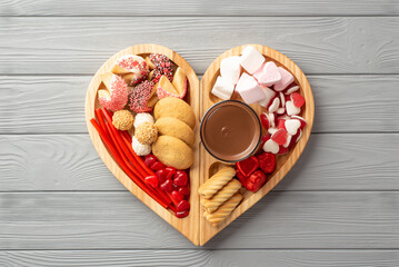 Valentine's Day concept. Top view photo of wooden heart shaped serving tray with confectionery candies cookies and glass cup of hot chocolate on grey wooden desk background
