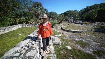 Mature woman wearing ethnic clothes, sunglasses, hat walking along the stone ruins of Xcambo Mayan pyramid in Yucatan Mexico. Concept of adventure is ageless.