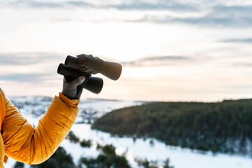 human hand in yellow jacket and warm glove holding binoculars againstwinter forest view of snowy river and sky Birdwatching ecology Research in nature, observation of animals Ornithology copy space