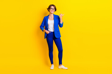 Fototapeta na wymiar Full length photo of positive shiny woman wear blue jacket spectacles pointing thumb empty space isolated yellow color background