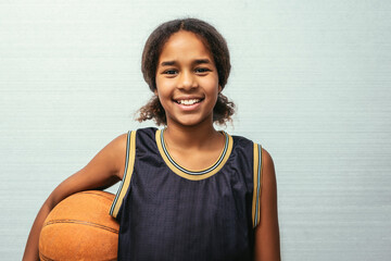 Female young basketball player standing against wall. Confident teenage girl is holding ball. She is wearing black jersey.