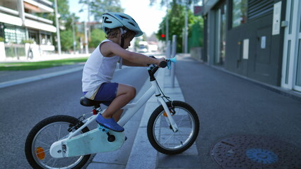 Fototapeta premium Little boy rides bicycle in city street. Back of child riding bike on sidewalk wearing protective helmet. Weekend childhood activity