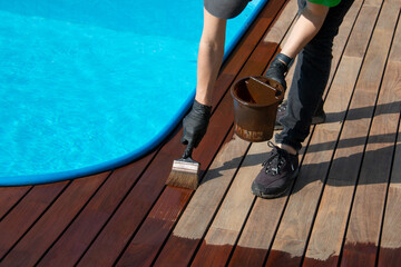 Worker applying brown wood protection oil on decking boards with paint brush next to the pool....
