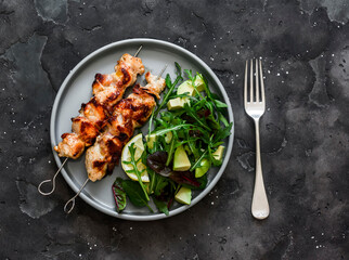 Chicken bacon skewers and  avocado, arugula, spinach, chard salad on a dark background top view