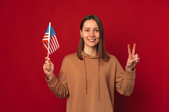 Cheerful Young Woman Is Holding Up USA Flag And Two Fingers Peace Gesture.