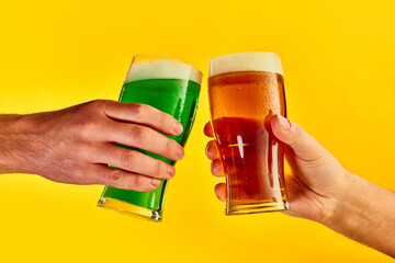 Cheers. Hands clinking green and lager foamy beer glasses over yellow background. Concept of st patrick's day celebration, brewery, traditions, alcohol drinks, taste, Irish holiday