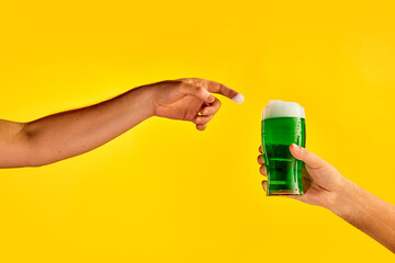 Male hand reaching glass with green foamy frothy beer over yellow background. Concept of st patrick's day celebration, brewery, traditions, alcohol drinks, taste, Irish holiday