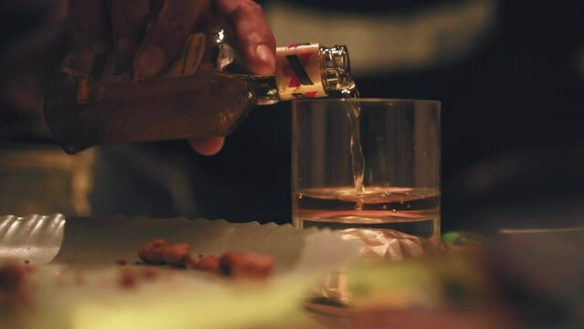 Close up of young adult male hand open bottle pours red wine transparent glass table indoor home. Unknown lonely man sitting alone picking up alcohol drink dark room house