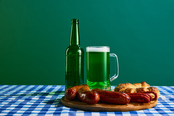 Mug with foamy green beer and plate of grilled sausages and buns on checkered tablecloth on green...