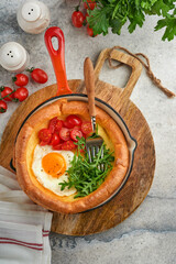 Dutch baby pancake. Fresh homemade Dutch Baby pancake with fried egg, tomato and green arugula in reed cast iron pan on light gray concrete rustic table. Flat lay, close up.