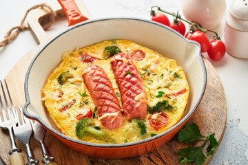 Omelette or frittata with two sausages, melted cheese, cherry tomatoes, broccoli and parsley on red skillet or iron pan on white old concrete background. Healthy breakfast food concept. Top view.