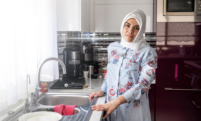 Middle-aged muslim woman does housework Kitchen cleaning sink wearing hijab headscarf. Islamic female washing home dishes. Housewife daily household life routine.