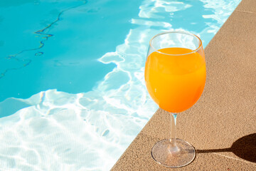 The orange juice in the wine glass is beside the swimming pool.
