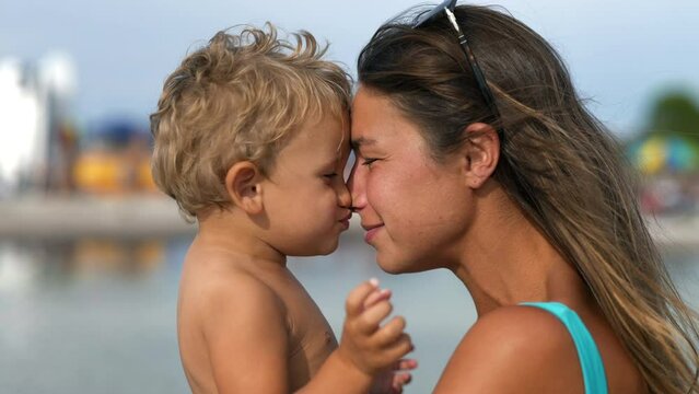 Portrait of Young Mother with her Toddler Enjoying Their Summer on the Beach. Loving and Affectionate Woman Laughing and Playing with her Kid  While Holding him in her Arms. Family on Vacation Concept