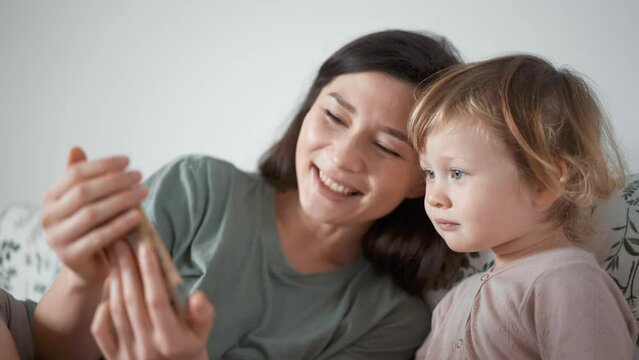 Asian young mother and curly boy daughter toddler kid look at phone screen video chat online. Woman and baby taking selfie or watch cartoons. Spend time together. Parenthood concept. bedroom morning