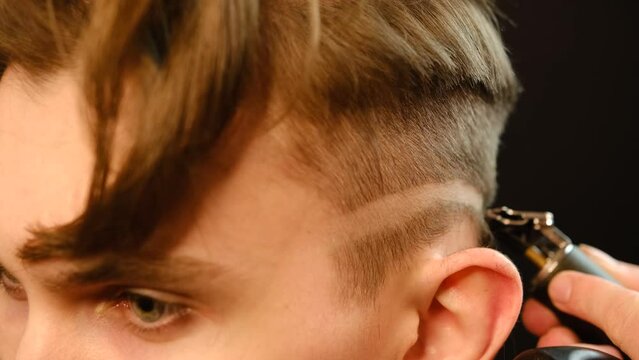 close up Shot of a handsome barber giving a haircut to his client using trimmer. Hairdresser service in a modern barbershop in a dark key lightning with warm light side view. Slow motion